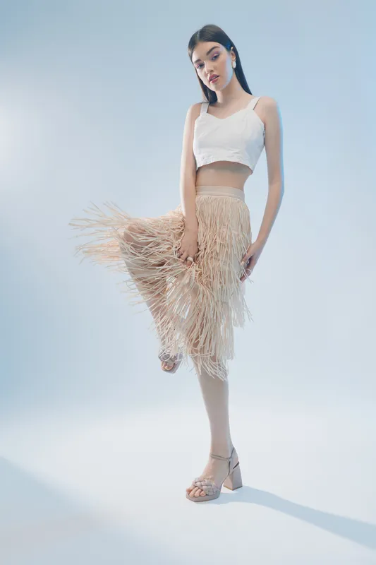 Young woman in a white crop top and beige fringe skirt poses elegantly on a light background. Concept of fashion, beauty, and modern style for editorial use.