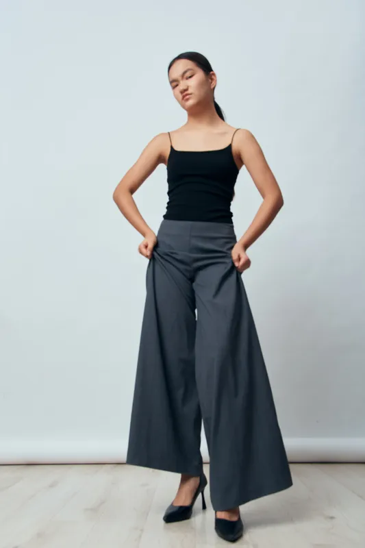 Young woman standing with hands in pockets wearing wide leg pants and black tank top on plain background. Casual fashion look with confident pose and minimal style indoors.