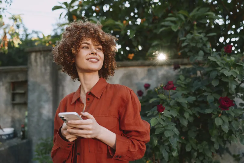 Confident woman with curly hair in a red shirt stands outdoors, holding a smartphone and smiling as sunlight blooms over a garden with roses
