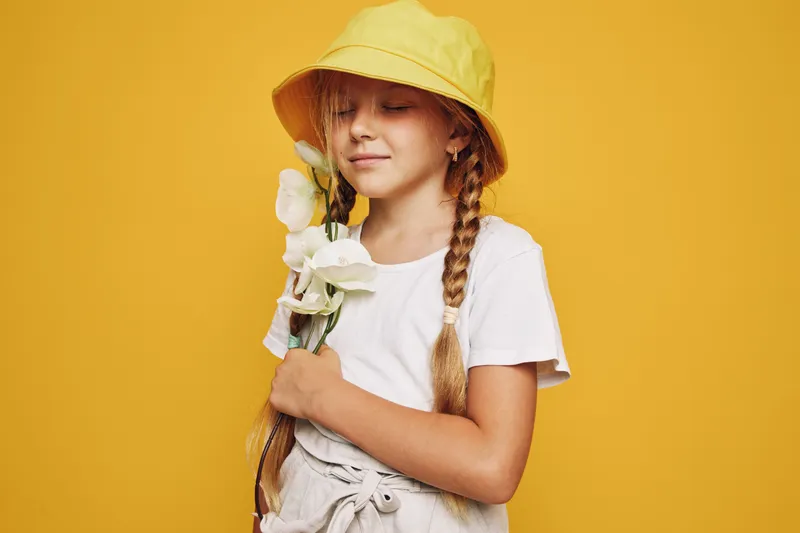 Young girl with braided hair wearing yellow bucket hat holds white flowers with closed eyes on yellow background. Concept of innocence and spring.