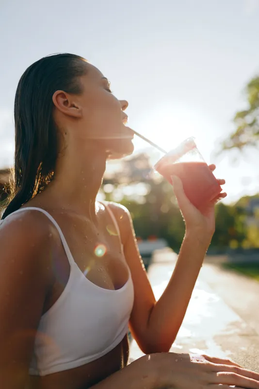 Healthy young woman drinks juice outdoors, basking in sunlight, wearing sleeveless white top, relaxed smile, summer vibe, casual lifestyle, isolated on bright street