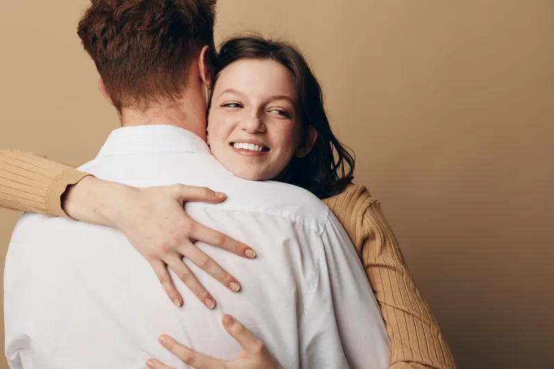 Happy young woman in beige sweater hugs man in white shirt, smiling with closed eyes. Intimate moment of affection, love, and warmth on neutral background, concept of relationship and trust.