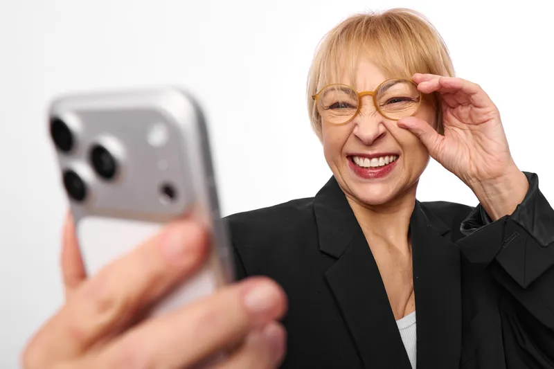 Smiling elderly woman adjusting glasses, holding smartphone for selfie, closeup with soft studio light, business blazer, technology lifestyle, communication and vision correction, warm mood