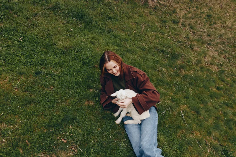 Woman lying on grass cradling a white goat kid, sharing a tender outdoor moment. Casual clothing, warm daylight, countryside calm, and gentle bonding in nature. soft textures, serene mood.