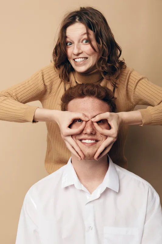 Smiling young woman in beige sweater playfully covers eyes of red haired man in white shirt. Studio portrait on beige background. Concept of friendship, fun, and joyful moments together.