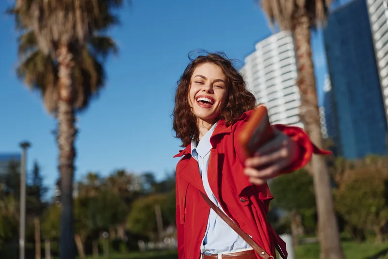 Young woman in a red coat smiles broadly while taking a cheerful selfie outdoors against a sunny urban backdrop, enjoying the spontaneous moment and vibrant street vibe.