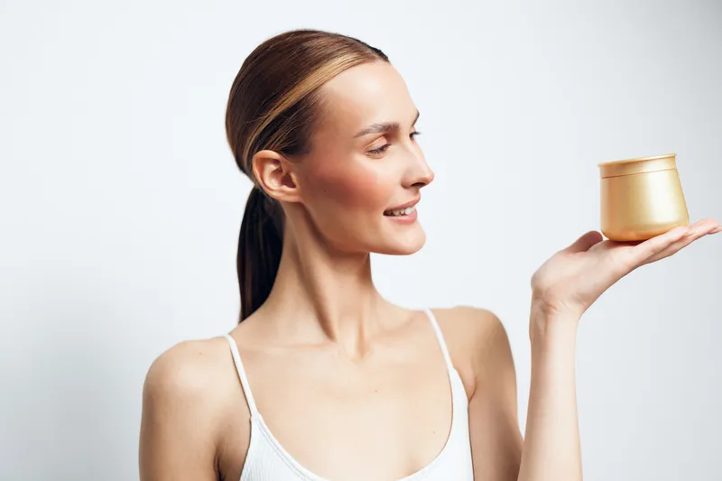 Smiling woman with a neat bun holds a small jar of cream, gazing at it with content. Close-up on a pale background. Skincare, wellness, and self care concept with soft studio lighting.