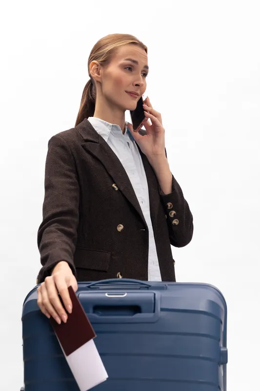 Businesswoman in dark jacket holds passport and boarding pass while talking on phone. Blue suitcase in foreground. Travel concept for business trip, communication, and planning.