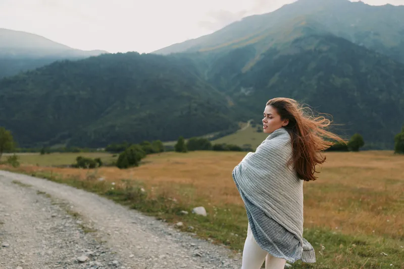 Woman standing on a dirt road in a valley wrapped in a light shawl looking to the side with wind in her hair Mountain view outdoor scene travel lifestyle calm mood