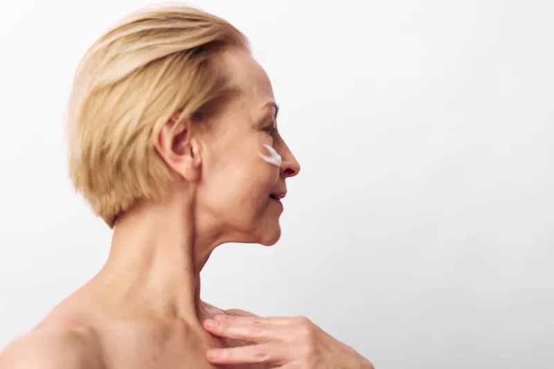 Mature woman with short blonde hair applies white face cream on cheek, showing skincare routine and healthy skin care concept with natural beauty on white background in soft lighting.