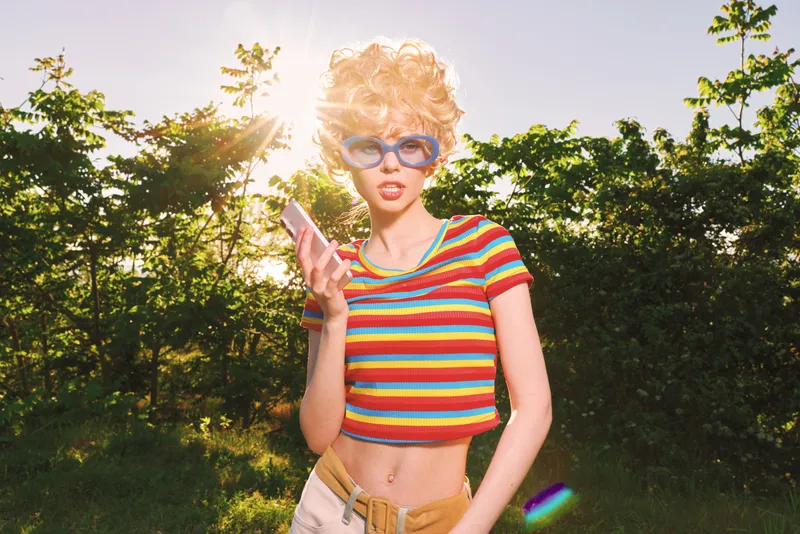 Smiling woman with curly hair wearing a striped crop top holds a smartphone outdoors, retro film colors, sun flare, casual lifestyle, candid fashion vibe on a warm day.