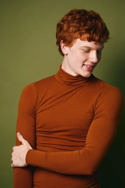 Young man with red hair wearing orange turtleneck sweater posing and smiling looking down against green background indoors portrait of casual fashion style.