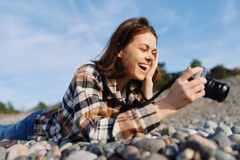 Woman lying on pebbles at the beach, capturing moments with a camera, wearing a plaid shirt, sunny outdoor scene, candid photography and joy.
