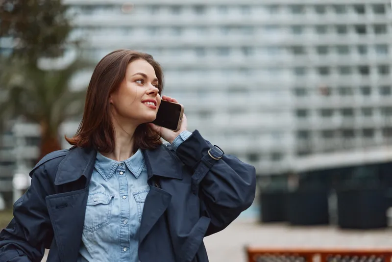 A smiling young woman in a blue coat talks on her smartphone outdoors near a modern building. Concept for communication, technology, and urban lifestyle.