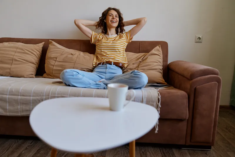 Smiling woman with curly hair relaxing on a sofa at home, casual striped shirt and jeans, cozy living space, warm natural light, lifestyle and comfort mood.