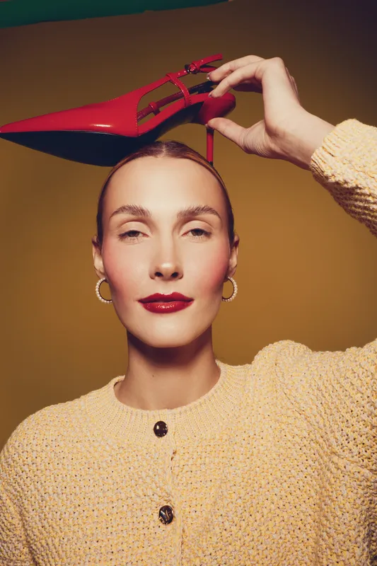 Woman with red lipstick and hoop earrings holds red high heel shoe on head. She wears beige knitted sweater against brown background. Concept of fashion, style, and elegance.