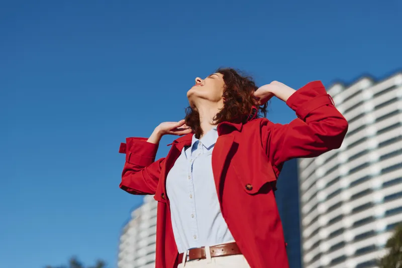 One young woman with curly hair in a red jacket poses outdoors, tilting her head back beneath a clear blue sky and modern buildings.