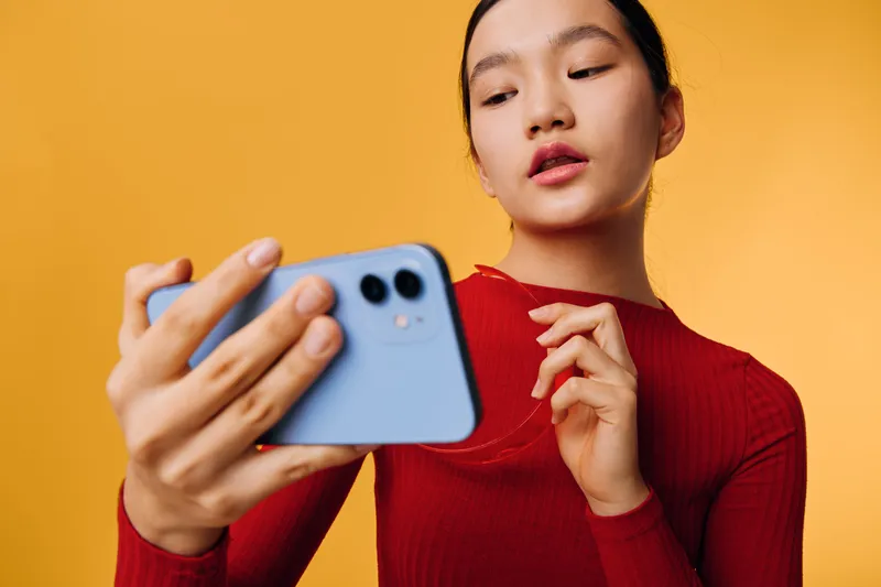 Young woman in red sweater taking selfie with smartphone against orange background, focused expression and casual pose with modern mobile device in hand, indoor portrait.