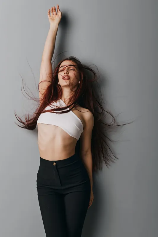 Young woman with long hair posing with raised arm against gray background. Casual outfit with white top and black pants, natural expression and movement in studio setting.