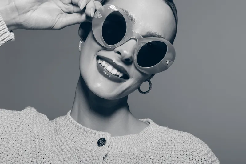 Smiling Caucasian woman with light skin wearing round sunglasses and knitted sweater poses happily. Close-up portrait with bright mood and casual fashion concept in studio lighting.