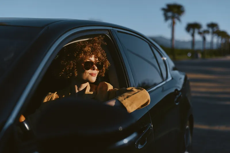Smiling woman with red curly hair driving a dark car, warm retro film colors illuminate the scene. Casual lifestyle and travel mood, a cheerful moment of freedom