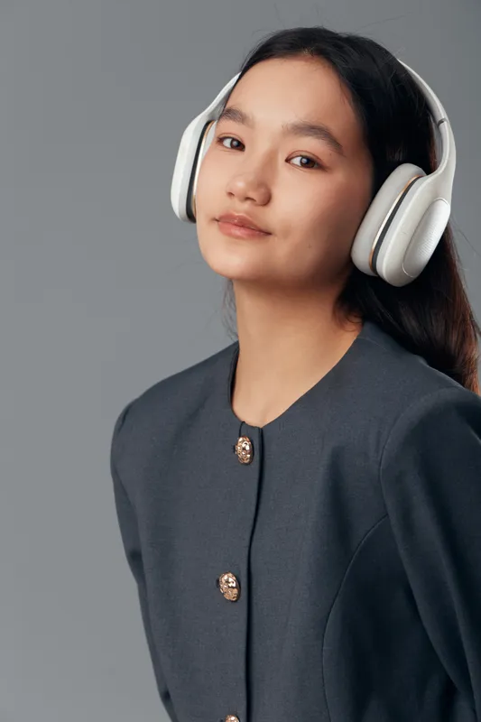 Young woman with long black hair wearing large white headphones and dark gray jacket posing against gray background. Portrait, technology, music accessory and modern style.