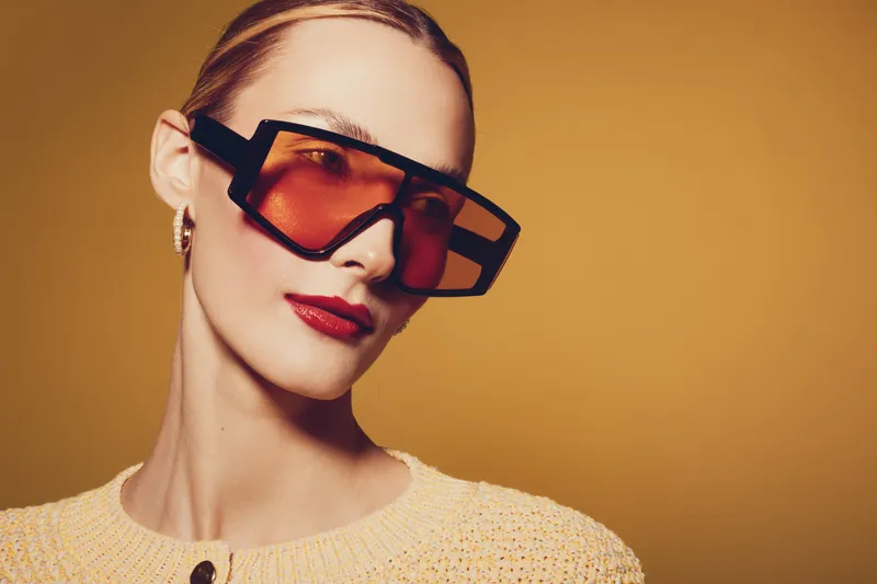 Stylish woman with slicked back hair wears oversized geometric sunglasses and red lipstick against a warm beige background. Fashion concept with modern accessories and makeup.