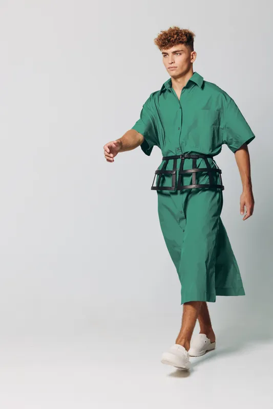 Young man walking in green oversized shirt and matching wide pants with black belt accessory and white sneakers on plain background in studio fashion portrait.