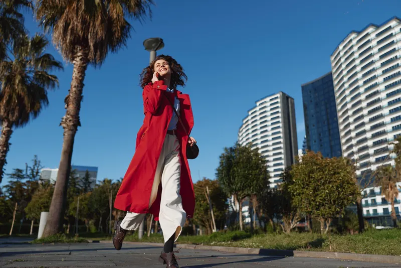 Young woman in a red coat walks confidently along an urban street lined with palm trees and modern buildings under clear blue sky.