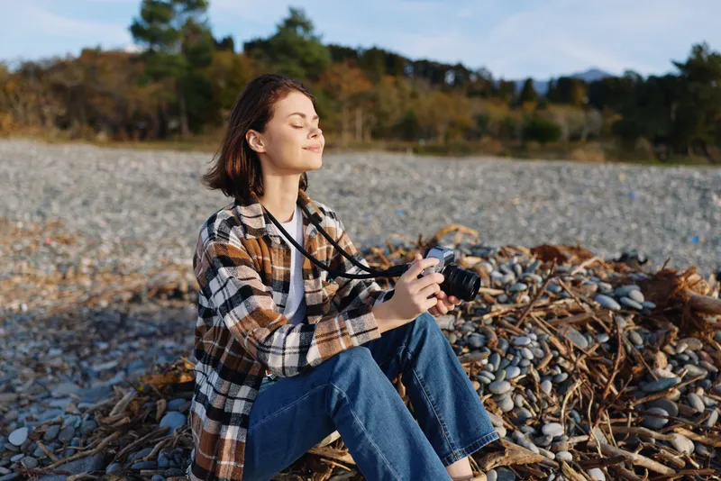 Woman photographer sitting on a rocky beach in a plaid shirt and jeans, enjoying sunny day while capturing coastal scenery with a camera and natural light.