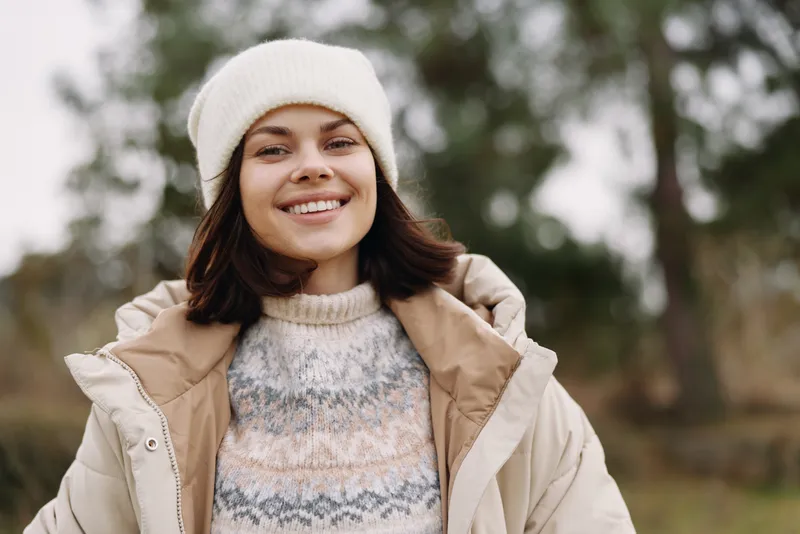 Woman outdoors in winter wearing a cozy beige coat and white beanie, smiling at the camera with blurred trees in the background, conveying seasonal warmth and cheerful style.