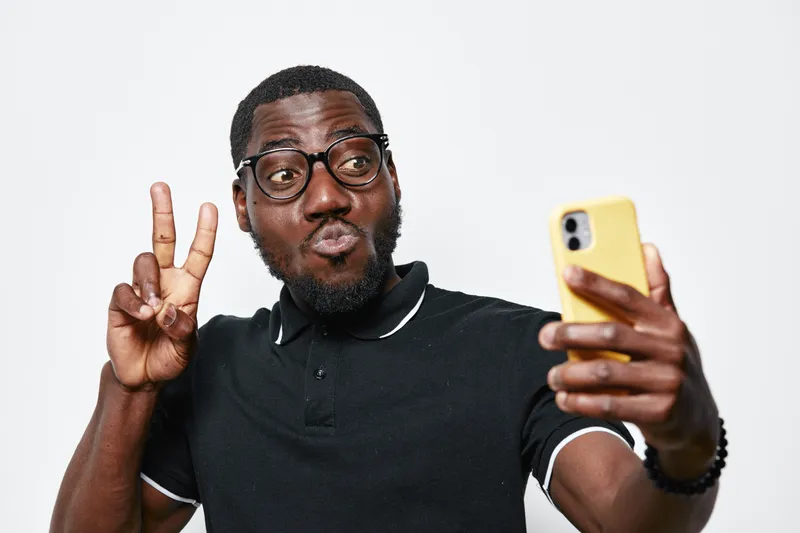 African American man with dark skin and glasses taking selfie with smartphone making peace sign wearing black polo shirt on white background.