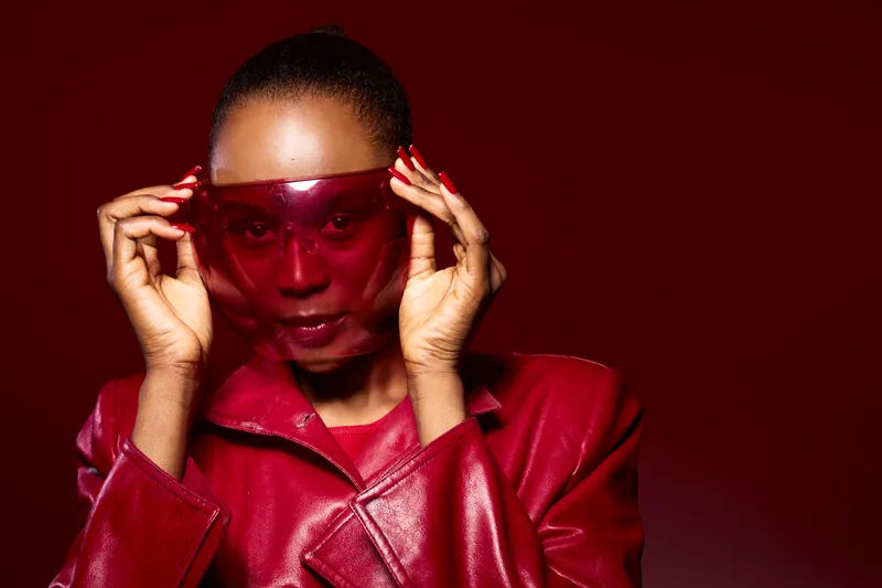 Young black woman wearing a red leather jacket and large red sunglasses adjusts eyewear against a dark red background. Concept of fashion, style, confidence, and modern beauty.