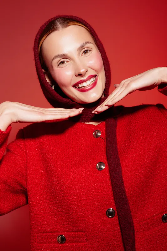Smiling woman in red coat and matching headscarf poses with hands near face on red background. Concept of fashion, winter style, happiness, and vibrant color contrast.