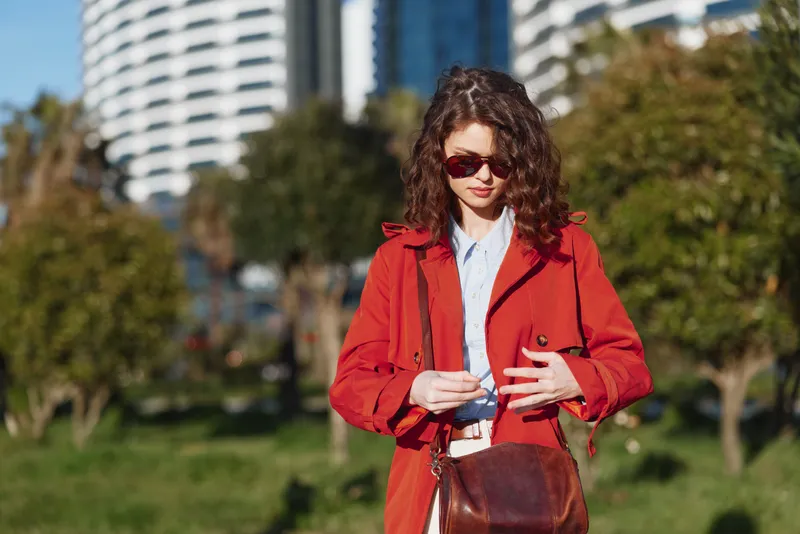 Young woman with curly hair wearing sunglasses and a red coat walking outdoors in a sunny urban park with modern buildings in the background.