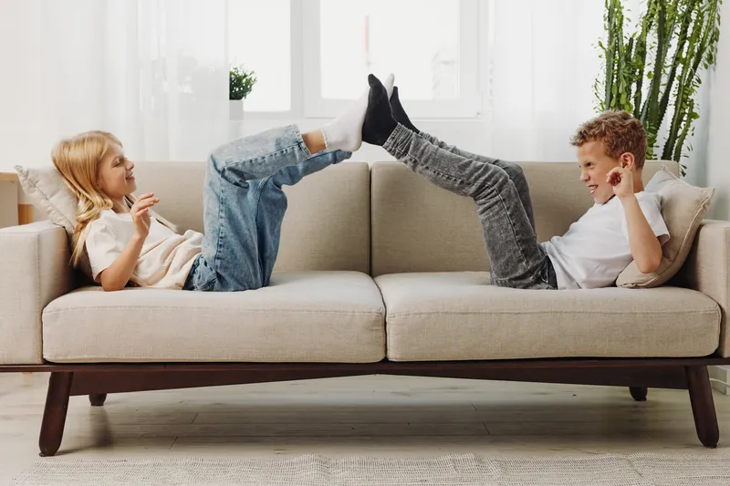 Two children lying on a grey sofa playfully touching feet and smiling, enjoying a fun moment in a bright living room with natural light and indoor plants. Concept of childhood joy and friendship.