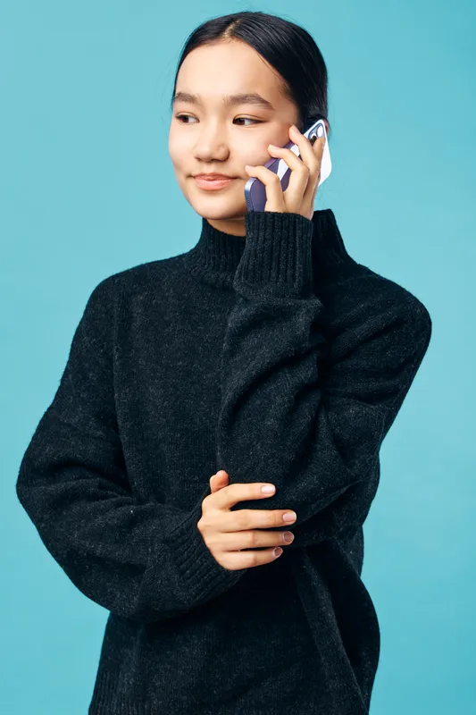 Young woman wearing black sweater talking on smartphone with relaxed smile against blue background casual communication technology modern lifestyle portrait