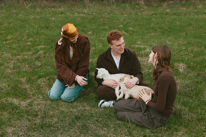 A small goat kid is gently petted by two women and a man outdoors, seated on grass in a park. A warm, casual moment capturing countryside lifestyle and animal companionship.