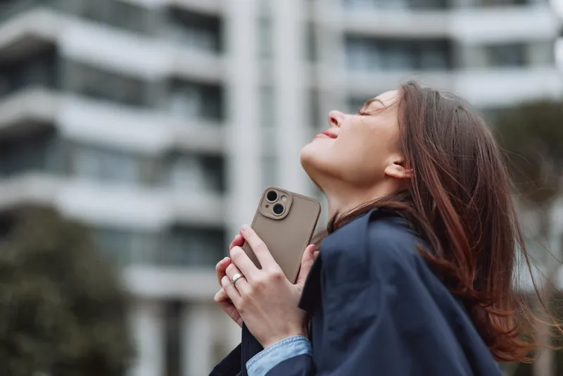 A brunette woman in a dark jacket raises her phone toward the sky, enjoying a moment of release in an urban outdoor setting.