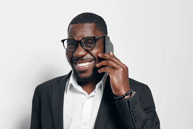 African American man with dark skin wearing glasses and black suit talking on smartphone smiling in studio with white background for communication and technology.