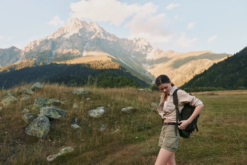 Young woman hiking in a sunlit alpine meadow with towering mountains behind, enjoying the fresh air and a relaxed moment