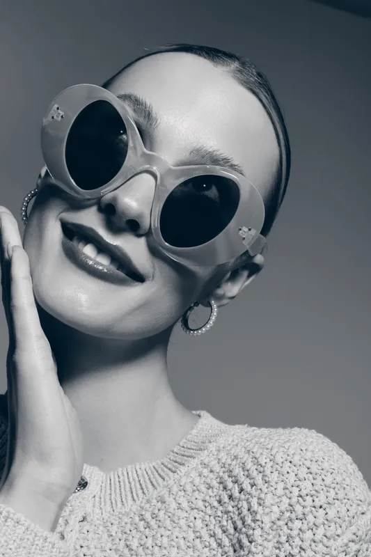 Portrait of a smiling white woman with pale skin wearing large round sunglasses and hoop earrings in a knitted sweater. Studio fashion concept with soft light.