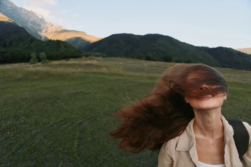 Young woman with long brown hair enjoys a windy day outdoors in a natural green field with mountains in the background, expressing freedom and relaxation.