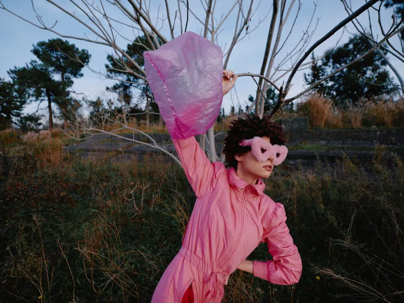 Woman in a pink dress raises a flowing scarf outdoors, dramatic pose with windy movement against a natural landscape, artistic fashion moment under open sky
