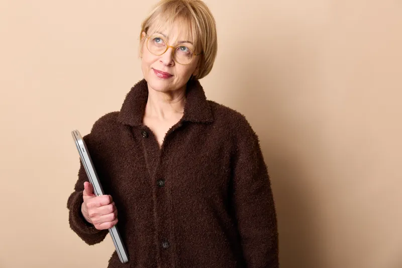 Woman, senior, glasses, tablet, technology studio portrait, warm brown fleece jacket, gentle smile, holding a tablet in hand, looking at camera, beige background, soft even light, calm indoor mood