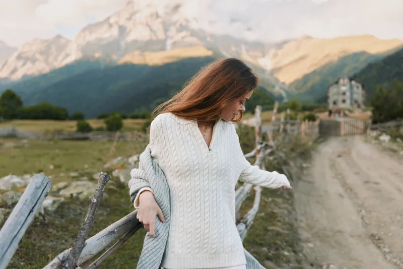 A young woman in a white knit sweater walks along a rural path with a wooden fence as mountains rise behind, conveying a calm, introspective lifestyle moment.