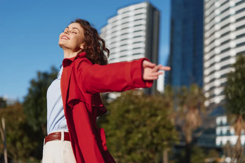 Young woman with curly hair enjoys fresh air outdoors wearing a red coat and white shirt with arms outstretched against a clear blue sky and urban background.