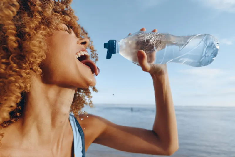 Young woman with curly hair drinking water from a plastic bottle outdoors near the sea, enjoying hydration on a sunny day with clear sky background.