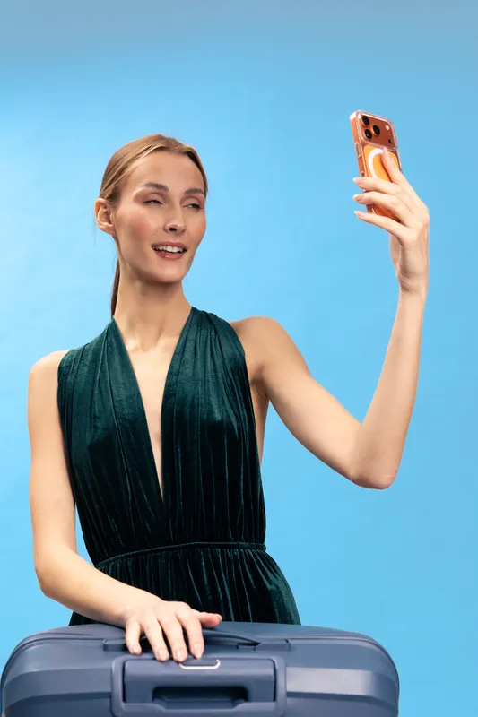 Young woman in dark dress taking selfie with smartphone while holding suitcase on blue background. Concept for travel, vacation, tourism, and modern lifestyle.