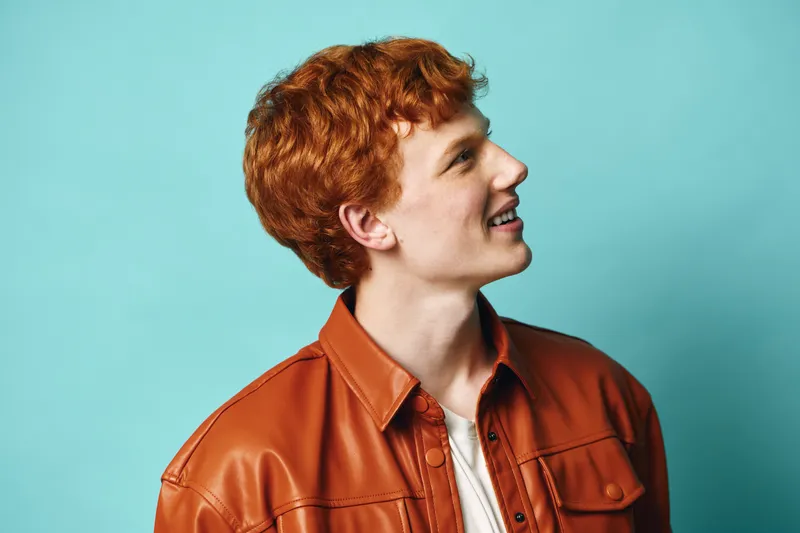 Young man with red hair wearing brown leather jacket posing in profile on blue background for fashion portrait and hairstyle concept.
